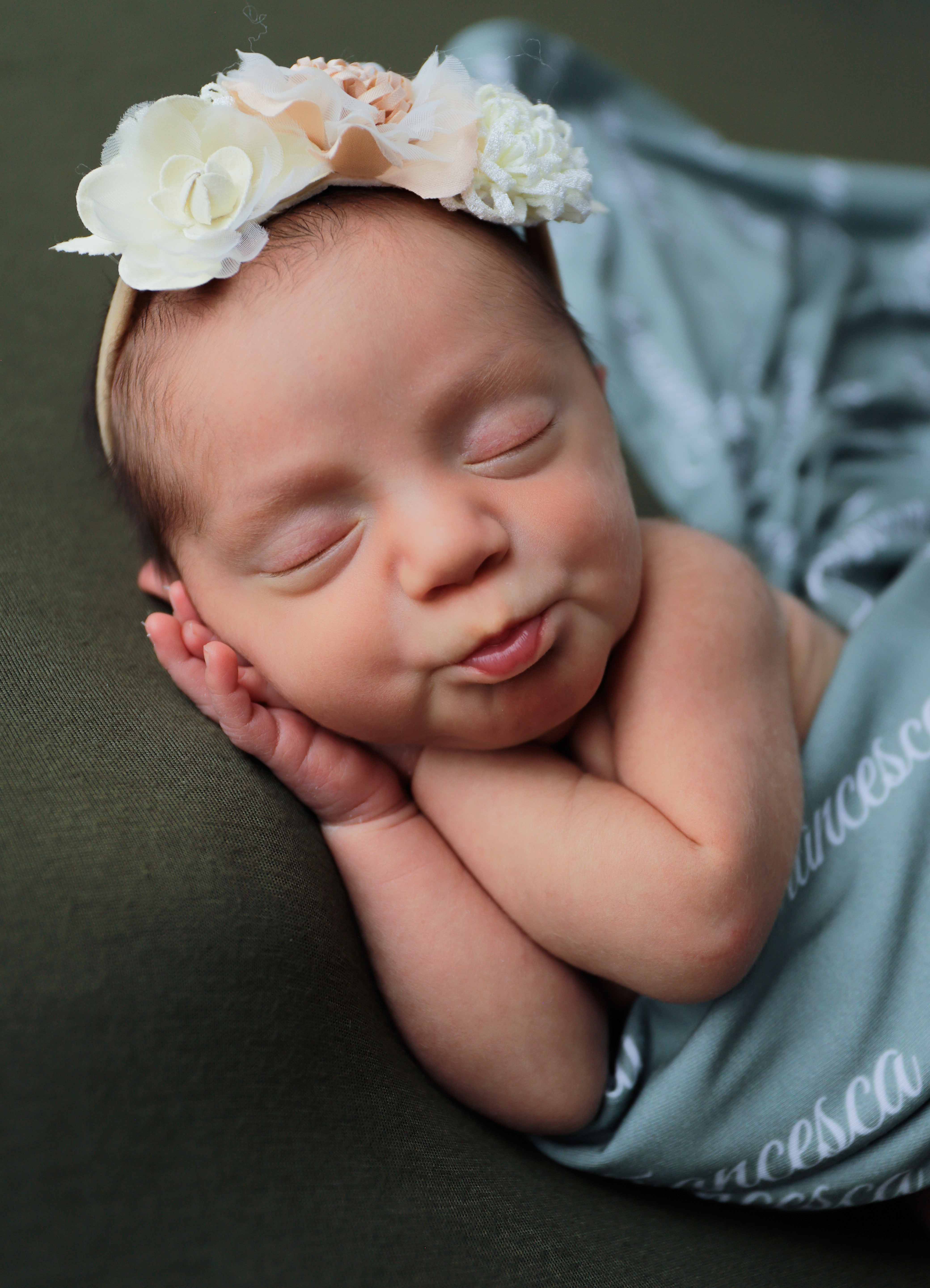 A newborn in soft taupe wrap, curled hands visible, sleeping.